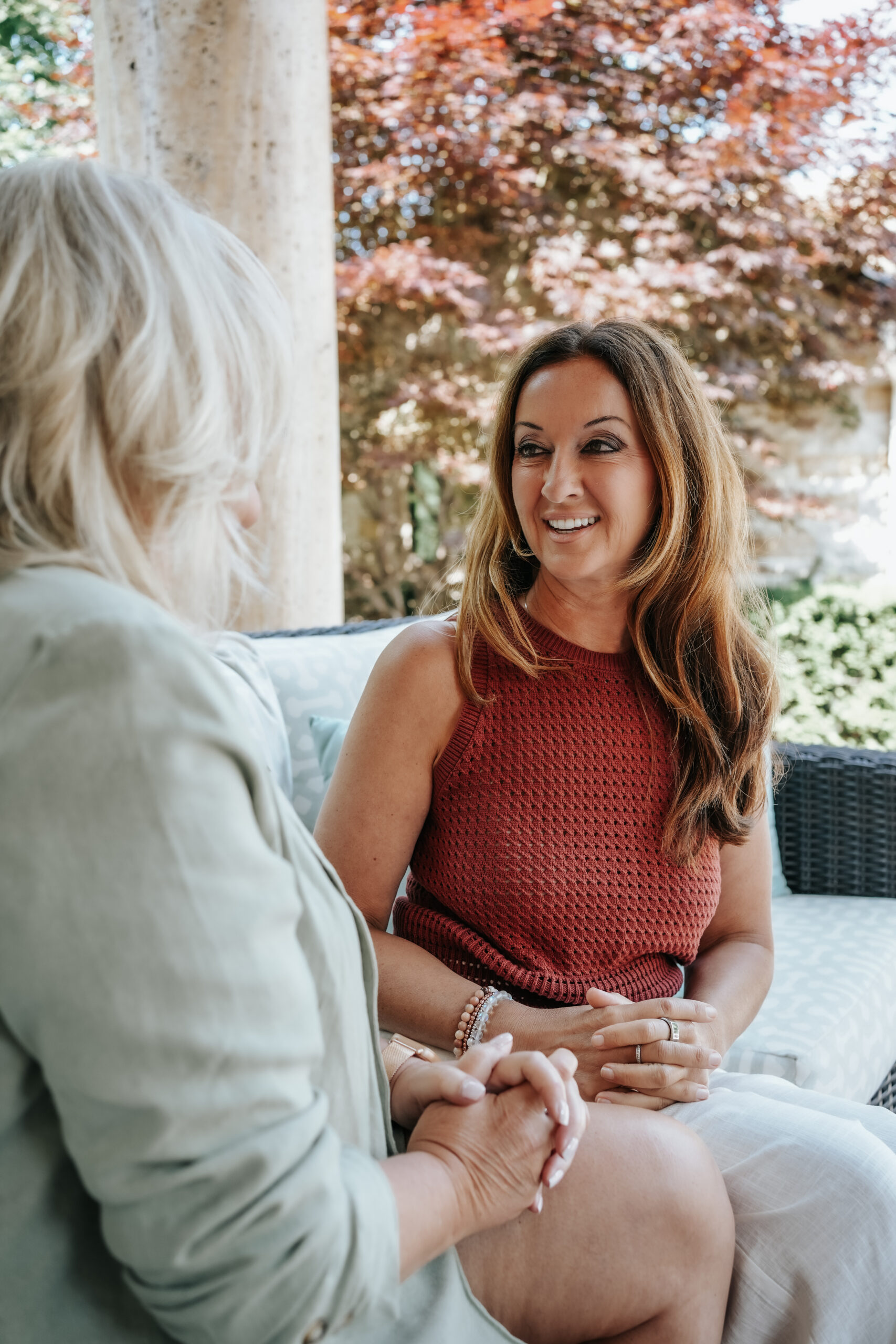 Kimberley Bertrand, Coach, sitting with a women and talk on a patio with trees in the background, one woman wearing a red sleeveless top and the other in a light-colored jacket.